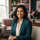 Portrait of a smiling South Asian woman in a professional setting with soft teal and rose-toned decor.