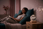 A South Asian woman relaxing in a dimly lit, rose and teal toned bedroom with a glowing 3D crystal lamp ball on a nightstand.